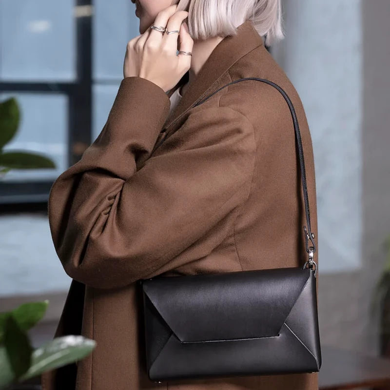 Schwarze Stella Umhängetasche aus Leder im Umschlag-Design, getragen über der Schulter mit einem braunen Wollmantel